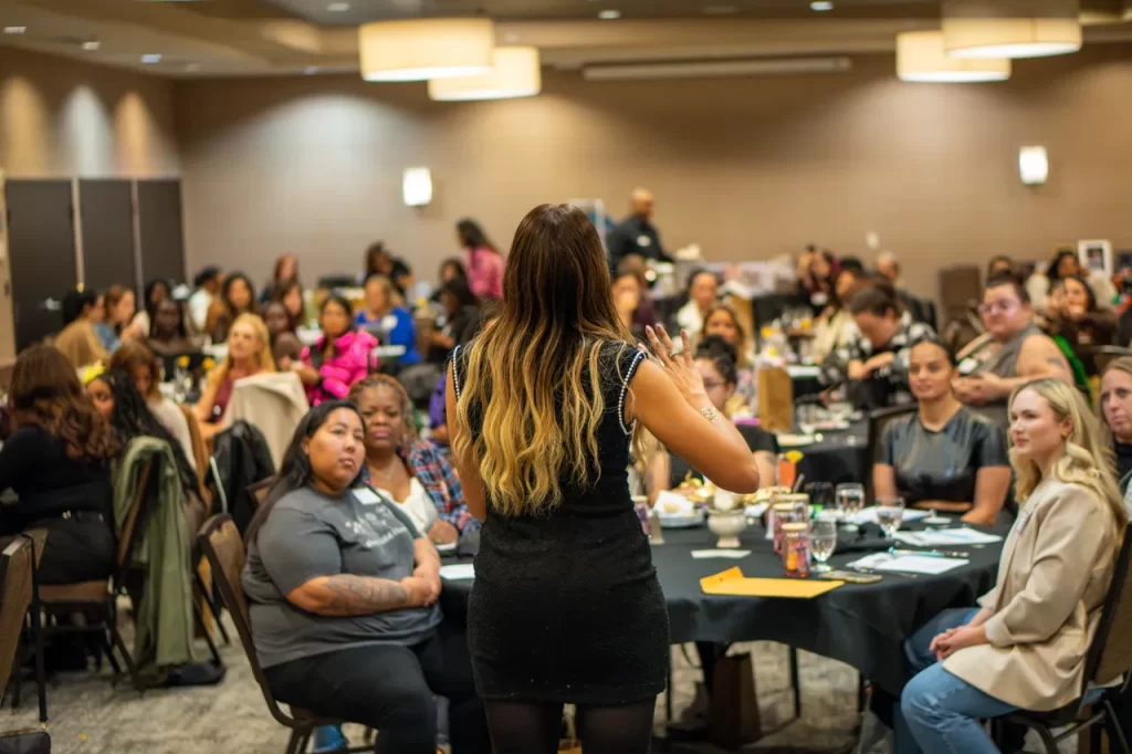 Moni stands and speaks, demonstrating her friendship coaching to an audience in a large conference room, fostering authentic relationships. People are seated at round tables, listening attentively. The room is well-lit, and theres a mixture of casual and formal attire among attendees.