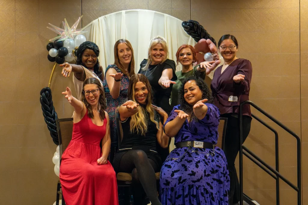 Moni and a group of seven other reliable friends, smiling and extending their arms towards the camera in an inviting gesture after completing the Winning In Friendship class. Embracing authentic relationships, they’re dressed in colorful outfits, seated and standing before a decorative arch adorned with balloons.
