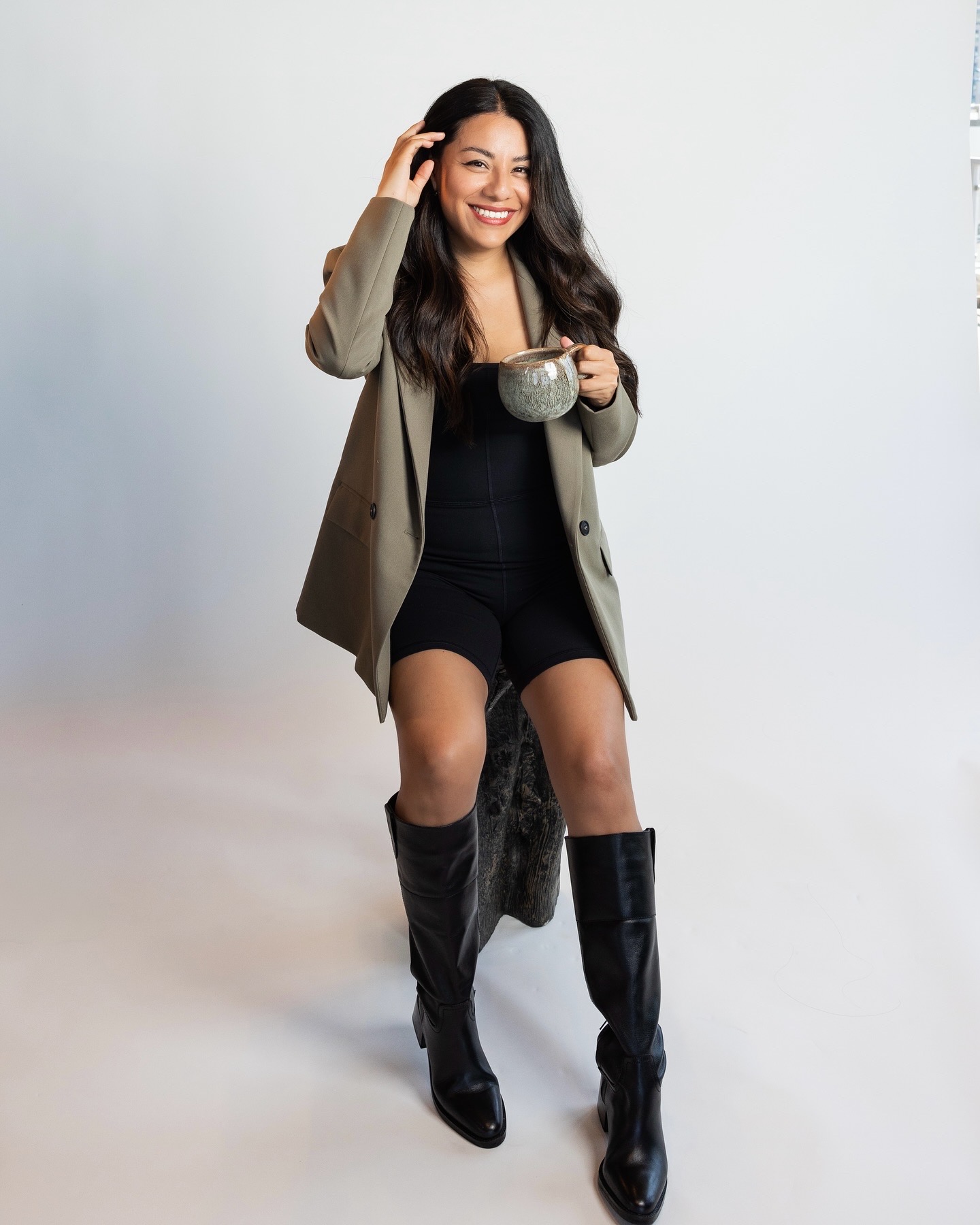 A woman in a black outfit and olive blazer sits on a stool, holding a cup and smiling, with one hand touching her hair. She is wearing knee-high black boots.
