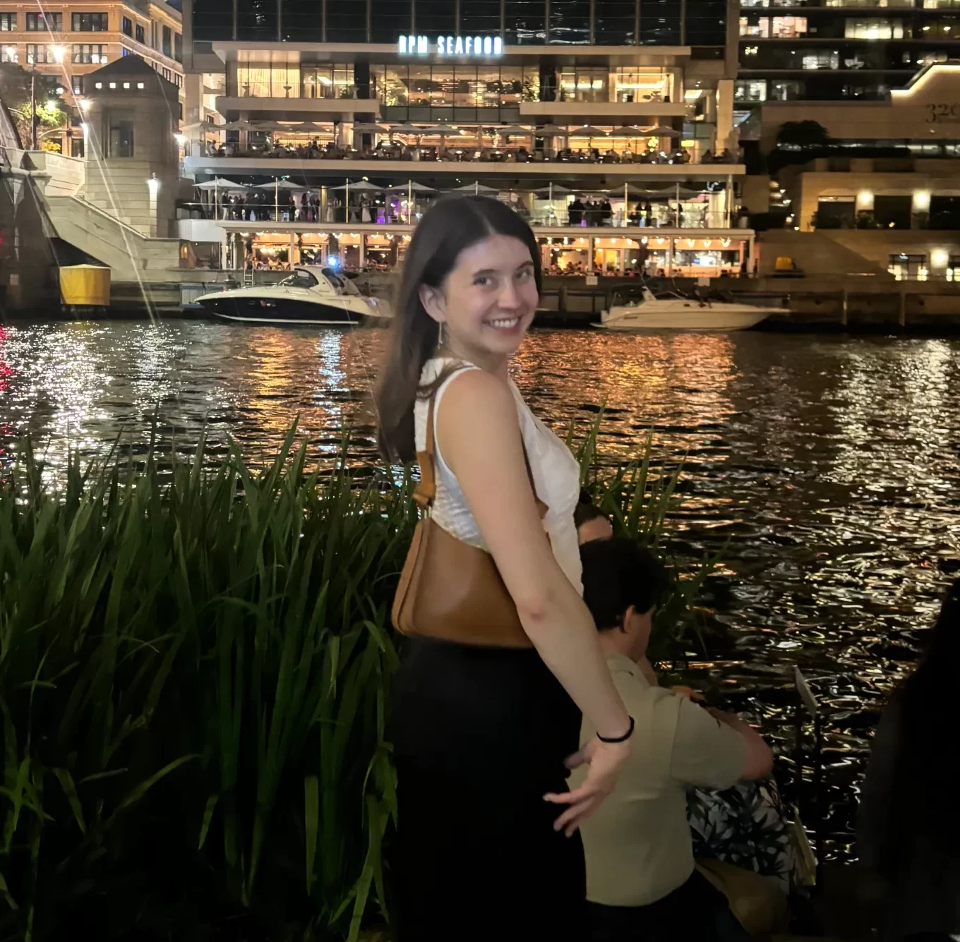A woman stands smiling by a riverside at night with buildings lit up in the background and other people seated near the water.
