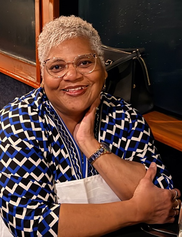 A woman with short gray hair and glasses smiles while seated, wearing a blue, black, and white patterned top and a watch, resting her head on her hand.