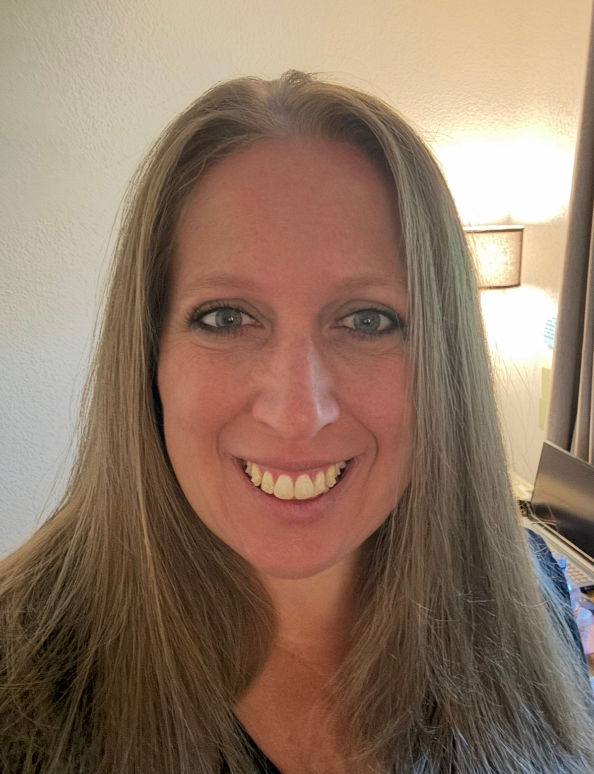 A woman with straight, light brown hair and blue eyes smiles at the camera in a well-lit indoor setting.