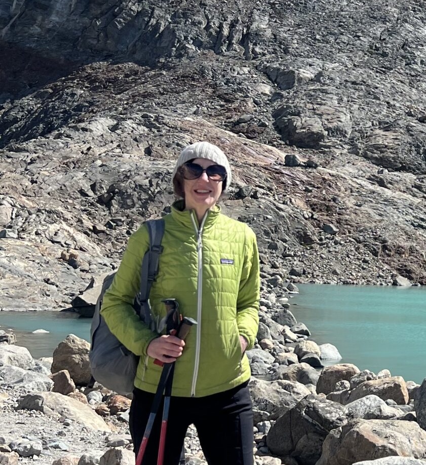 A person in a green jacket and knit hat stands on rocky terrain by turquoise water, holding trekking poles and carrying a backpack.